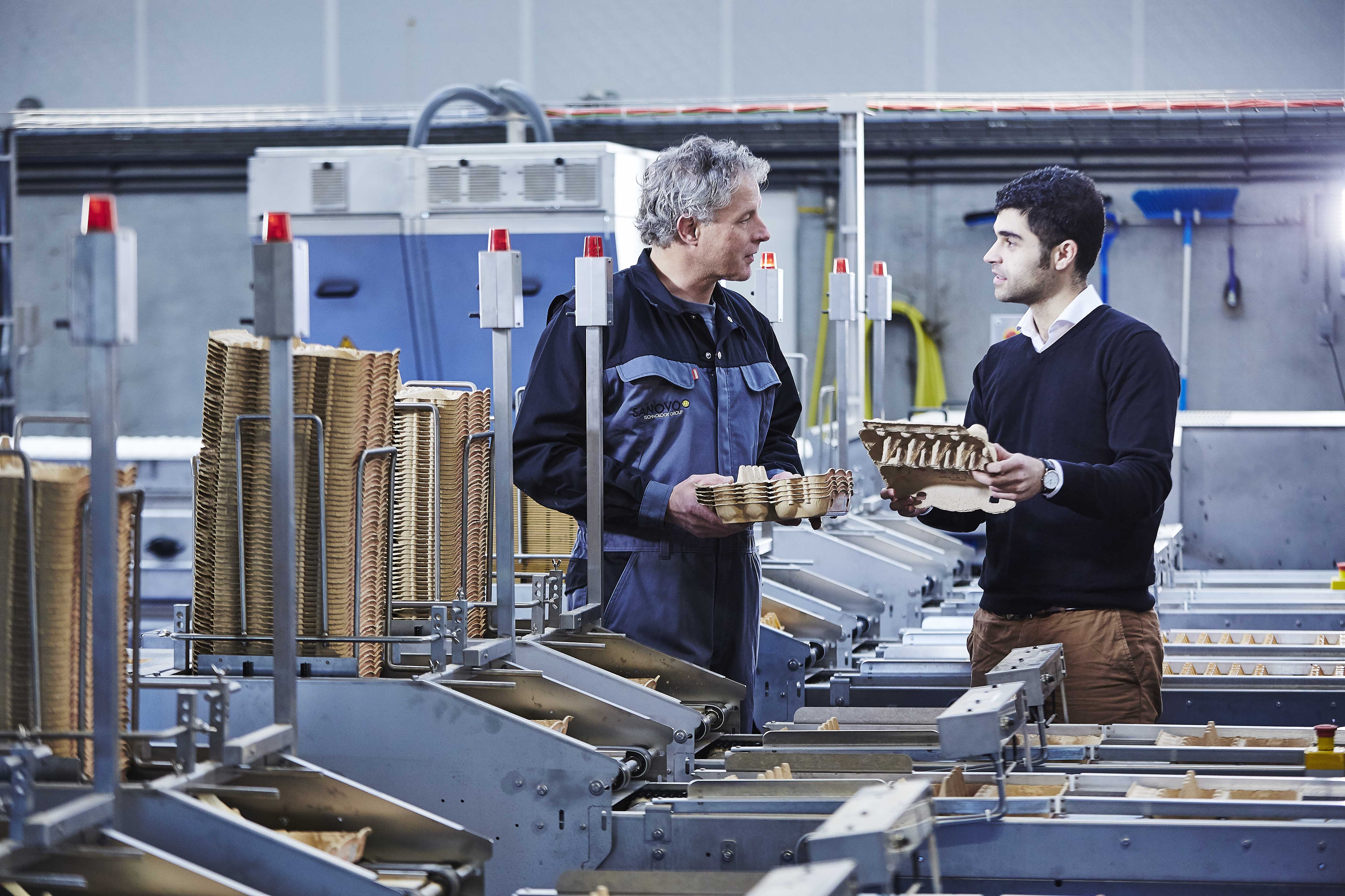 Two men holding egg trays and discussing next to egg-handling equipment.