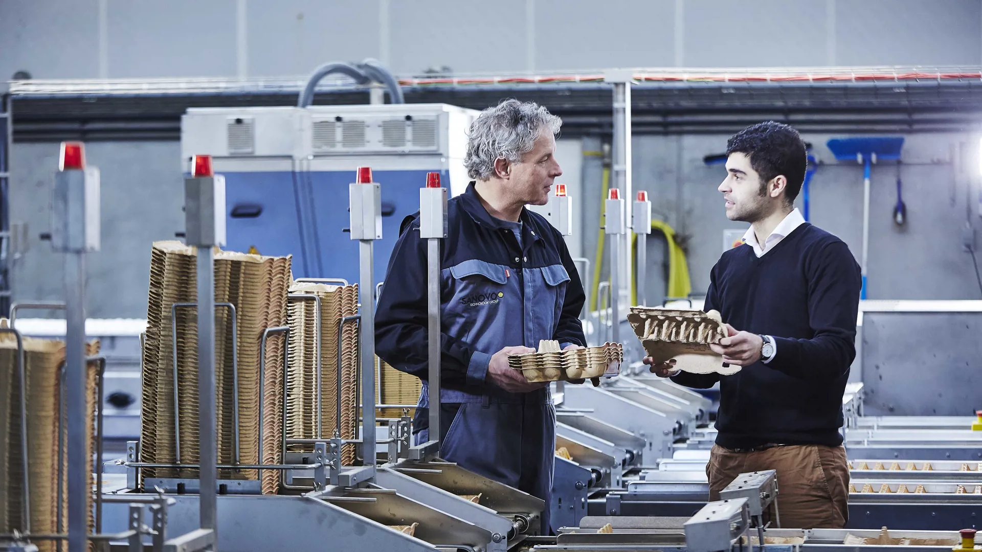 Two men holding egg trays and discussing next to egg-handling equipment.