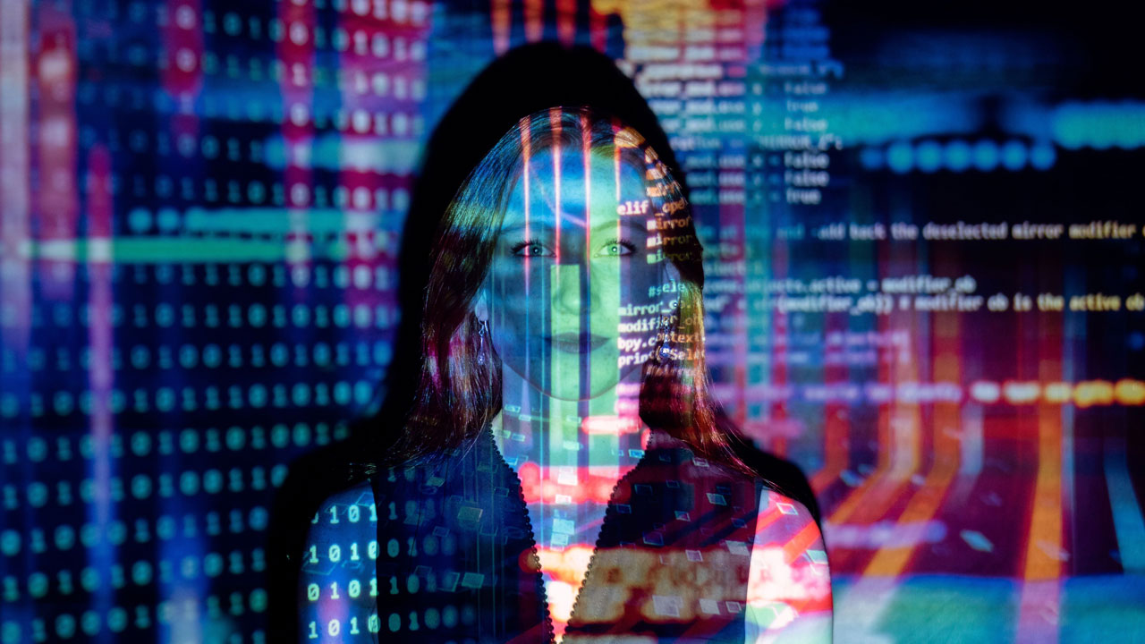 Woman standing in front of a digital screen with colorful data code and binary patterns projected onto her face and background.