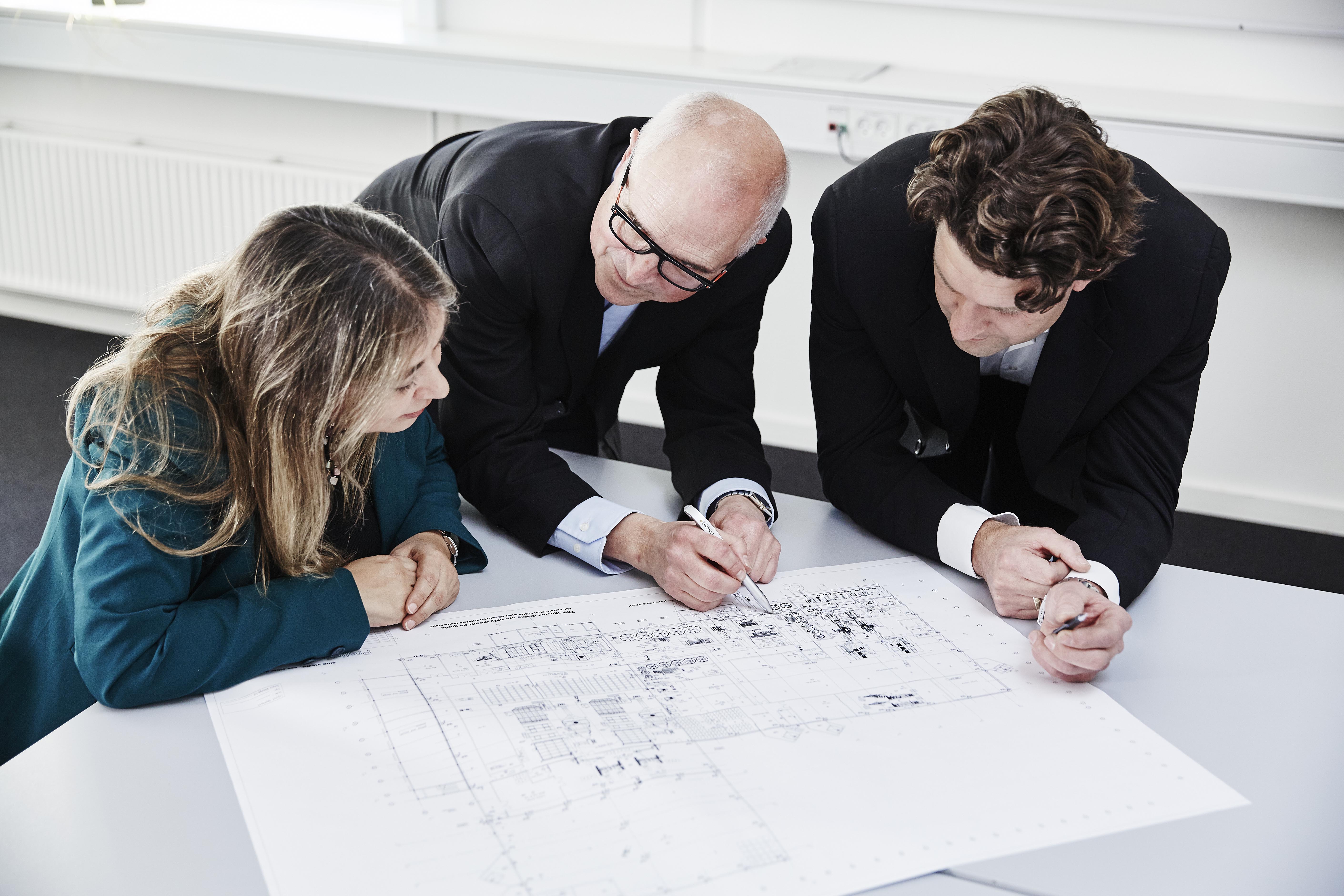 Three people reviewing and discussing a technical layout drawing at a table in an office environment.